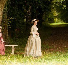 2 femmes en costume d'époque dans une jardin boisé : 1 assise sur un banc, l'autre debout