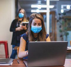 Une jeune lycéenne portant un masque fixe l'écran de son ordinateur posé sur un bureau en bois